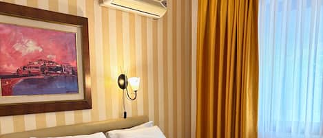 A hotel room with a bed, a nightstand, an air conditioning unit, a wall-mounted light, a painting, and a window with curtains.