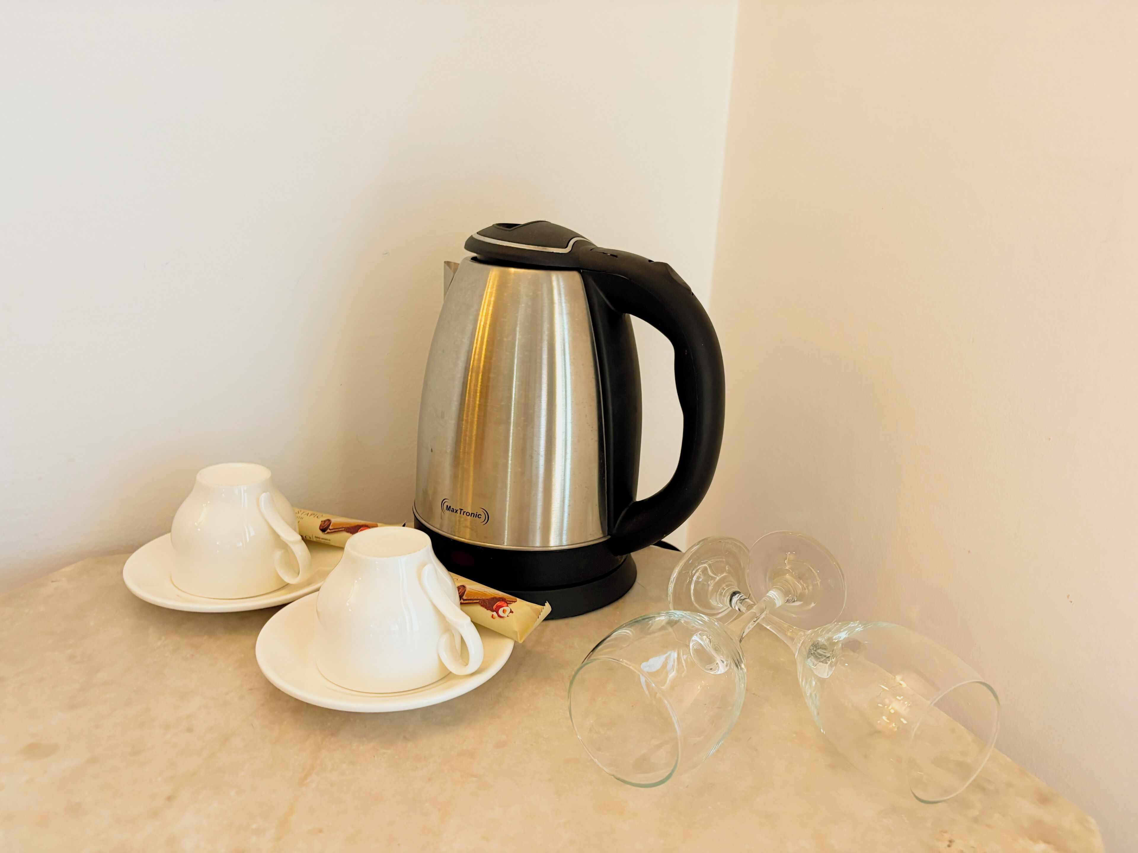 Una tetera de acero inoxidable con asa negra, dos tazas blancas con sus platillos y dos copas de vino transparentes sobre una superficie de mármol.
