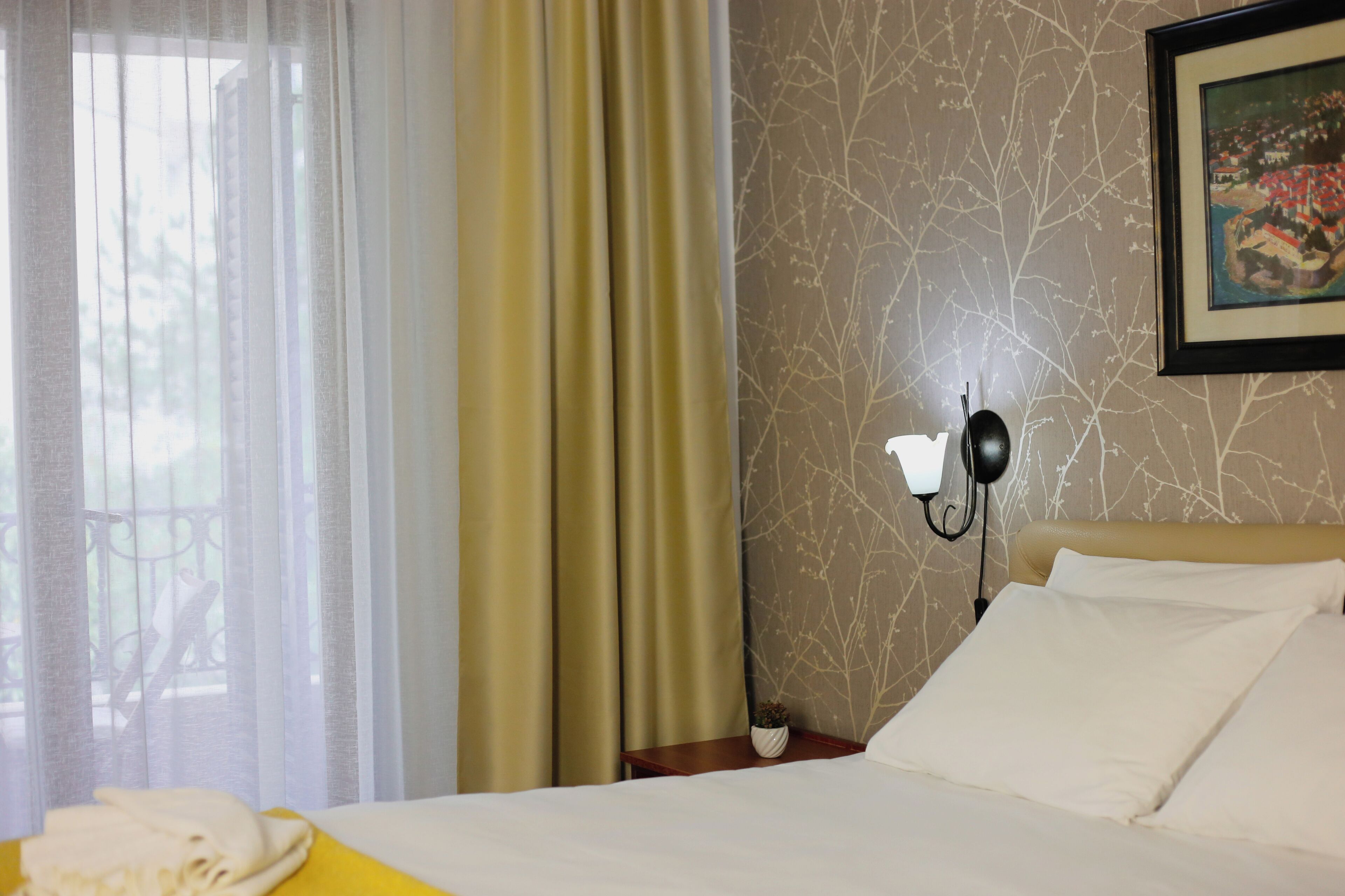 Una habitación de hotel con una cama, cortinas, una lámpara fijada a la pared y una pequeña mesa con una planta.