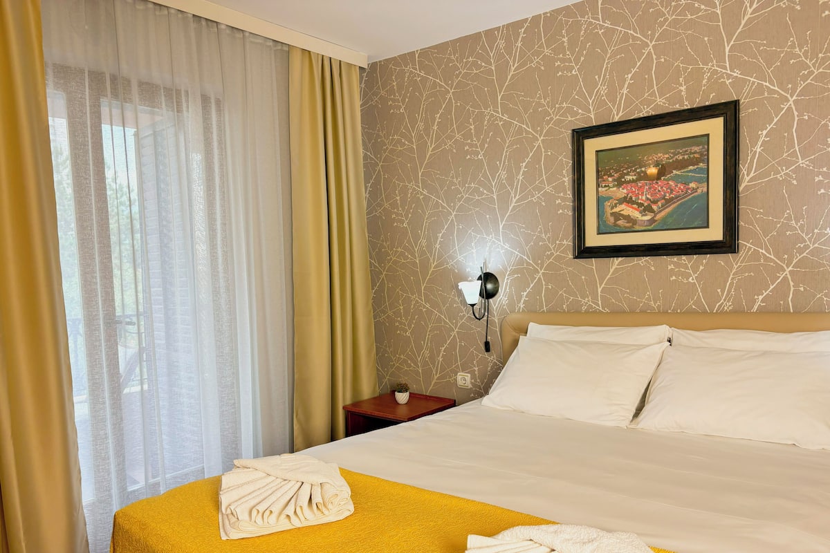 Une chambre d’hôtel avec un lit, une couvre-lit jaune, des oreillers blancs, une table de chevet avec une lampe et une fenêtre avec des rideaux légers.