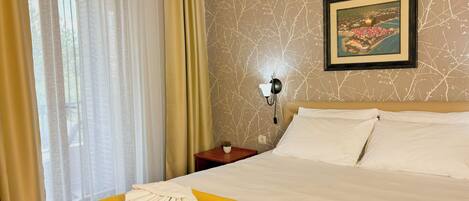 A hotel room with a bed, yellow bedspread, white pillows, a nightstand with a lamp, and a window with sheer curtains.