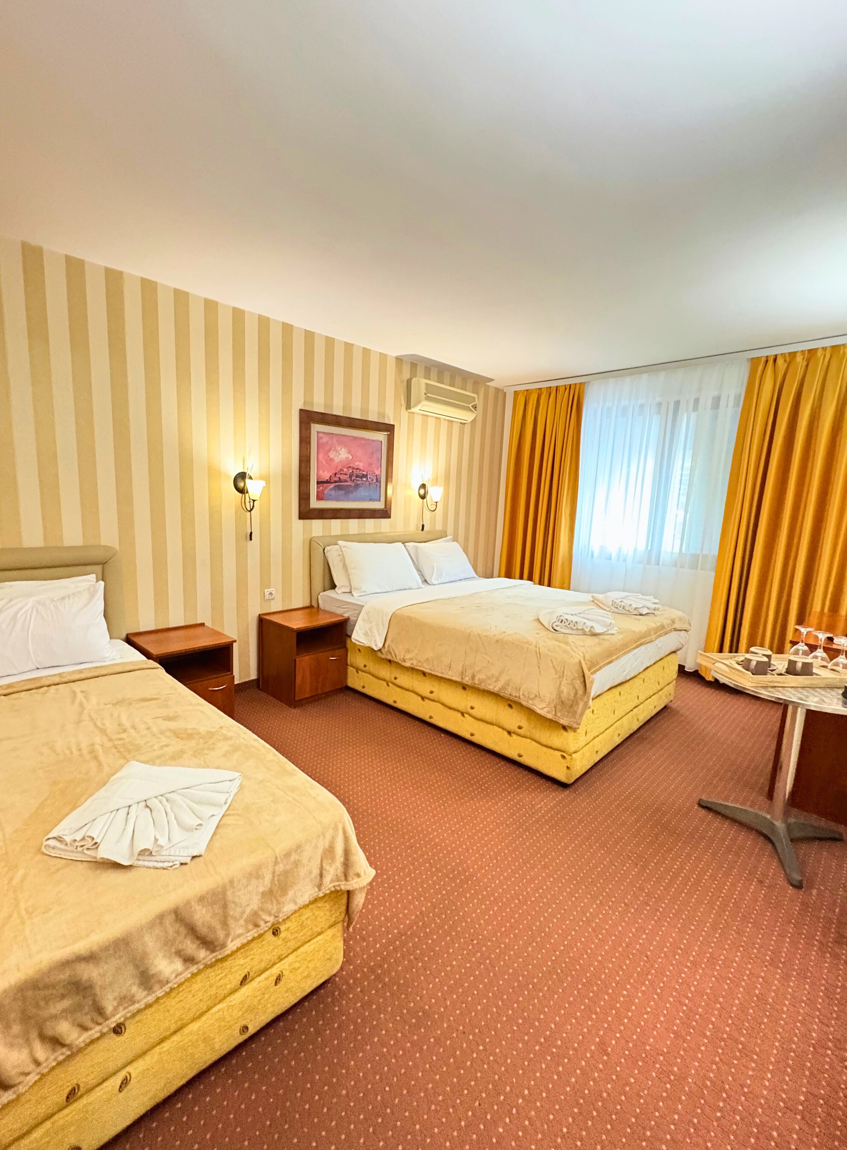 A hotel room with two beds, striped wallpaper, a framed picture, an air conditioner, and a table with a tray.