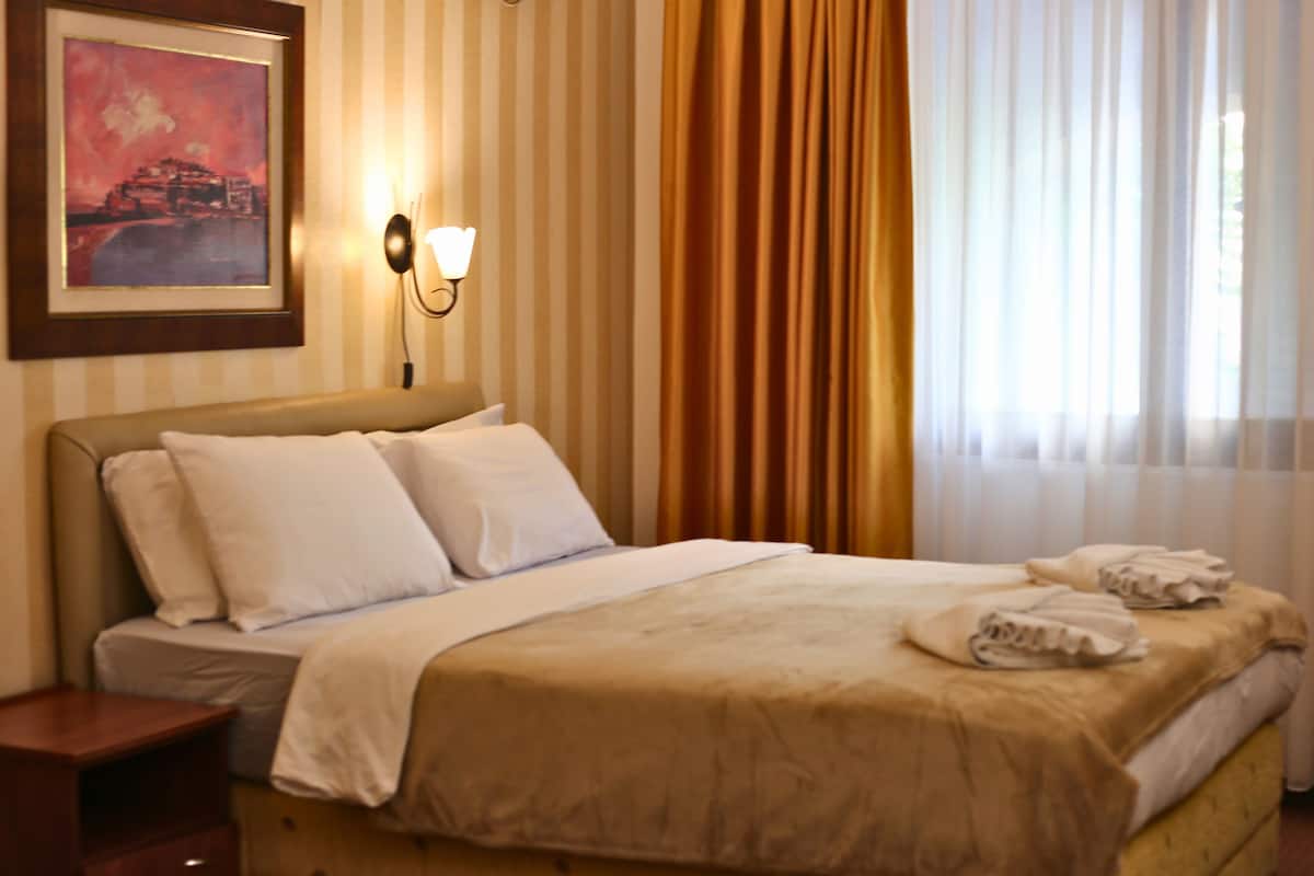 Une chambre d’hôtel avec un lit, une table de chevet, une lampe fixée au mur, un tableau encadré et une fenêtre avec des rideaux.