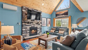 A cozy living room with a stone fireplace, a ceiling fan, and a large window. - Luxury 3-Story Pocono Retreat w/ Sauna+Hot Tub+Games (Albrightsville)