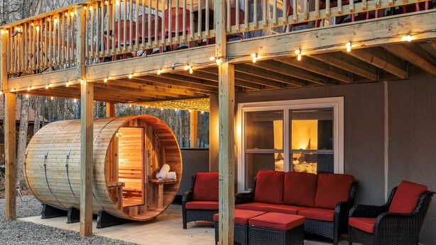 A wooden deck with string lights, a large wooden barrel, wicker furniture, and a glass door leading to a room. - Luxury 3-Story Pocono Retreat w/ Sauna+Hot Tub+Games (Albrightsville)