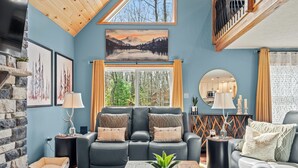 A modern living room with a stone fireplace, a large window with curtains, a black leather sofa, and a wooden coffee table. - Luxury 3-Story Pocono Retreat w/ Sauna+Hot Tub+Games (Albrightsville)
