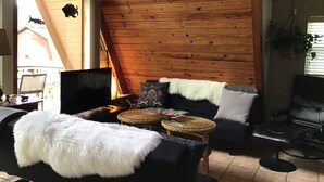 A cozy living room with a wooden ceiling, a black leather sofa, a white furry throw, and a television.