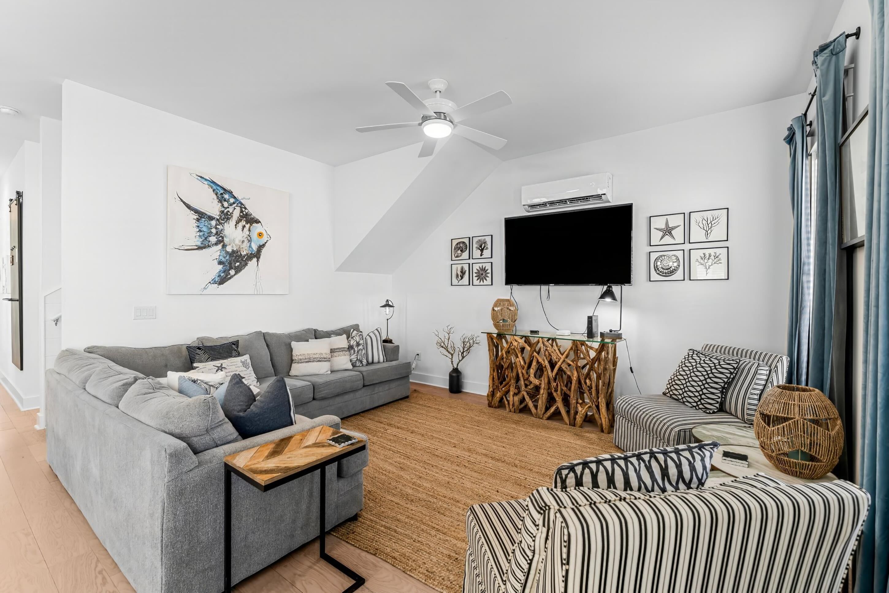 A modern living room with a grey sofa, a striped ottoman, a wooden coffee table, a ceiling fan, and a flat-screen TV mounted on the wall.