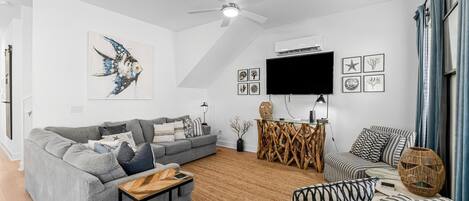 A modern living room with a grey sofa, a striped ottoman, a wooden coffee table, a ceiling fan, and a flat-screen TV mounted on the wall.