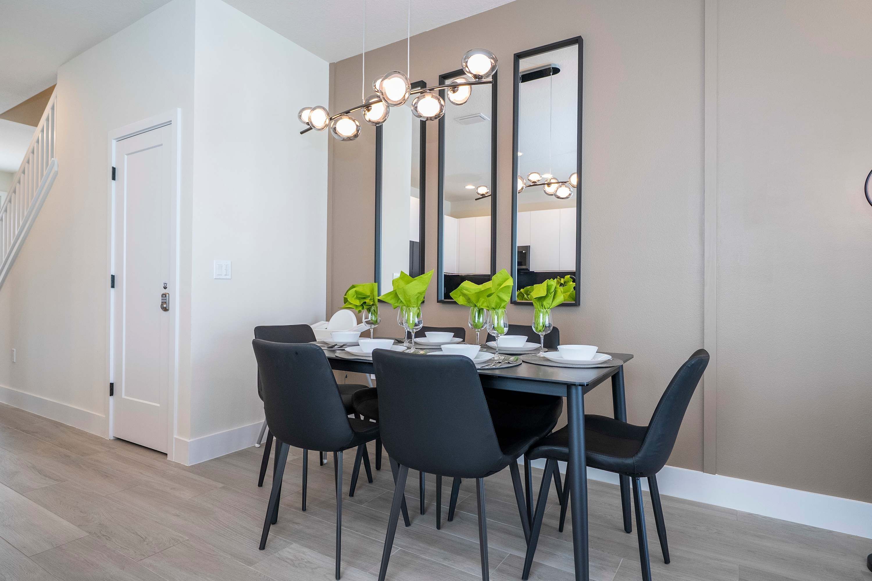 Un comedor con una mesa dispuesta para cuatro personas, sillas negras, una araña de cristal y un espejo grande en la pared.