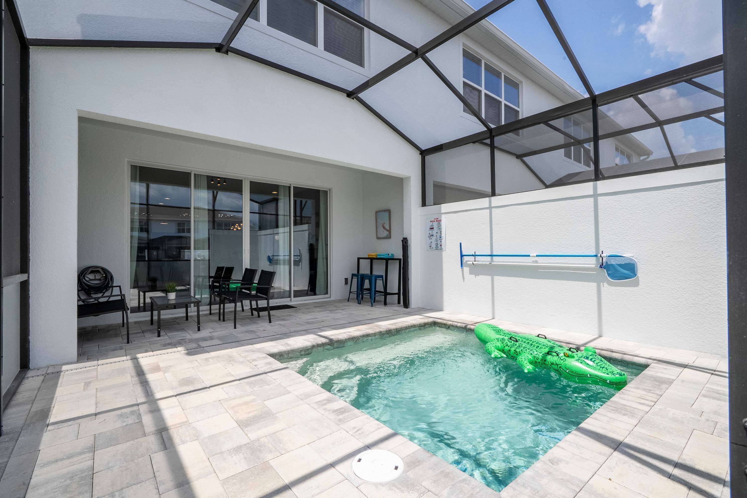 A swimming pool with a green inflatable crocodile, a covered patio area with chairs and a table, and a glass door leading to a room with a view.