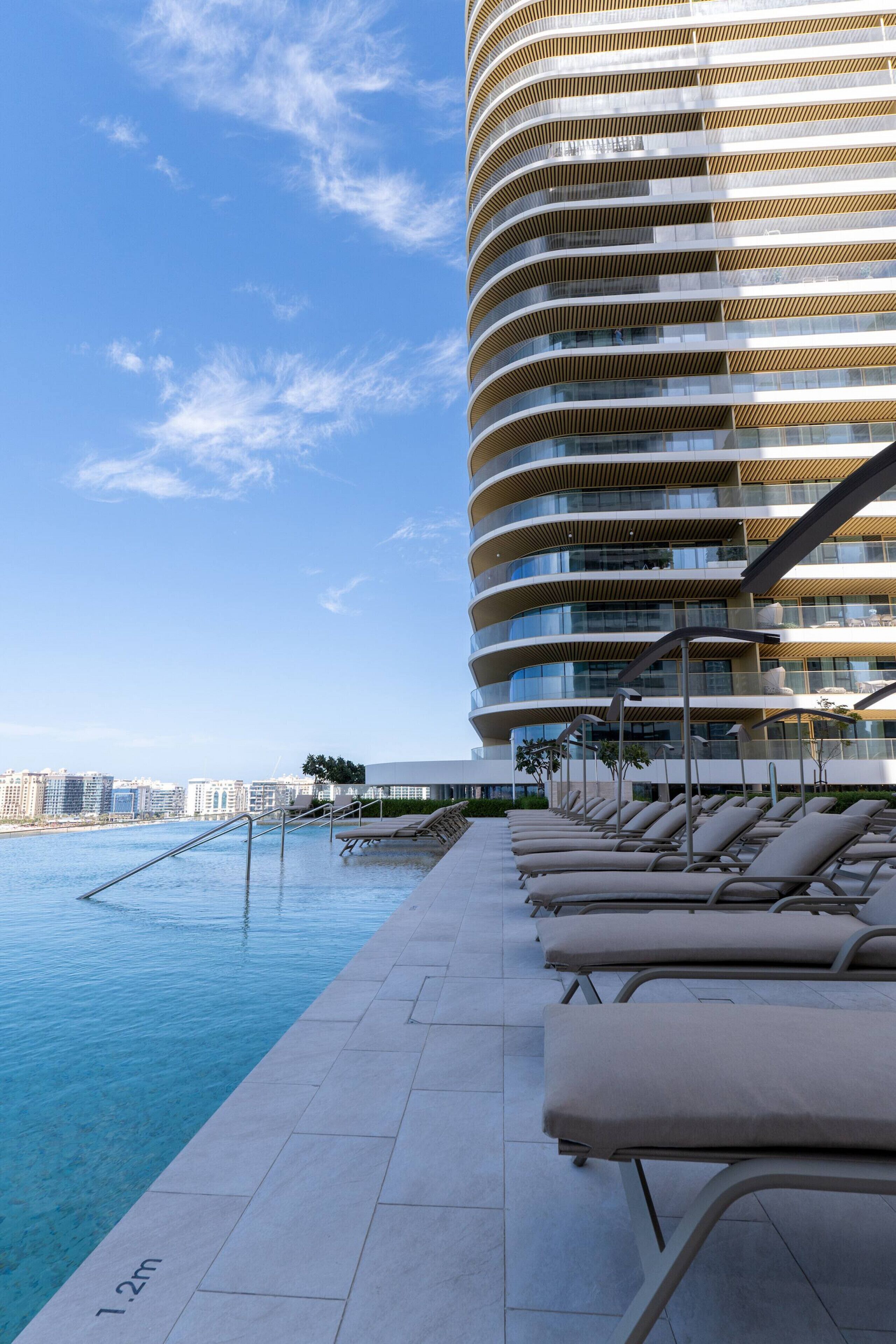 Un hotel moderno con facciata curva, area piscina con poltrone relax e vista chiara sullo skyline della città sullo sfondo.
