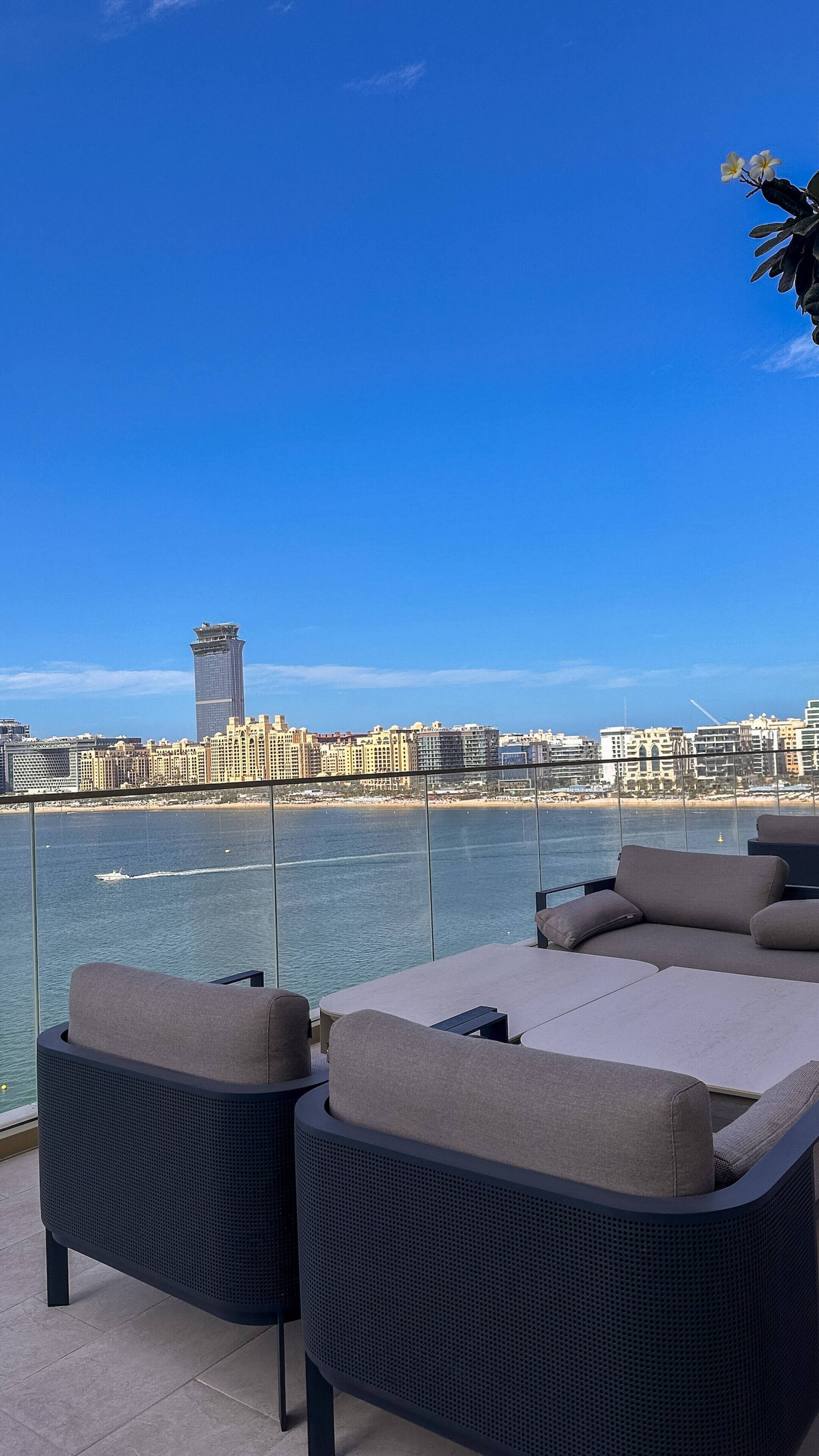 A balcony with two wicker lounge chairs and a glass railing overlooking a cityscape and water.