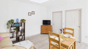 A living room with a sofa, a wooden dining table set for two, a television on a wooden cabinet, and a ceiling light. - Bathach Beag (Oban)