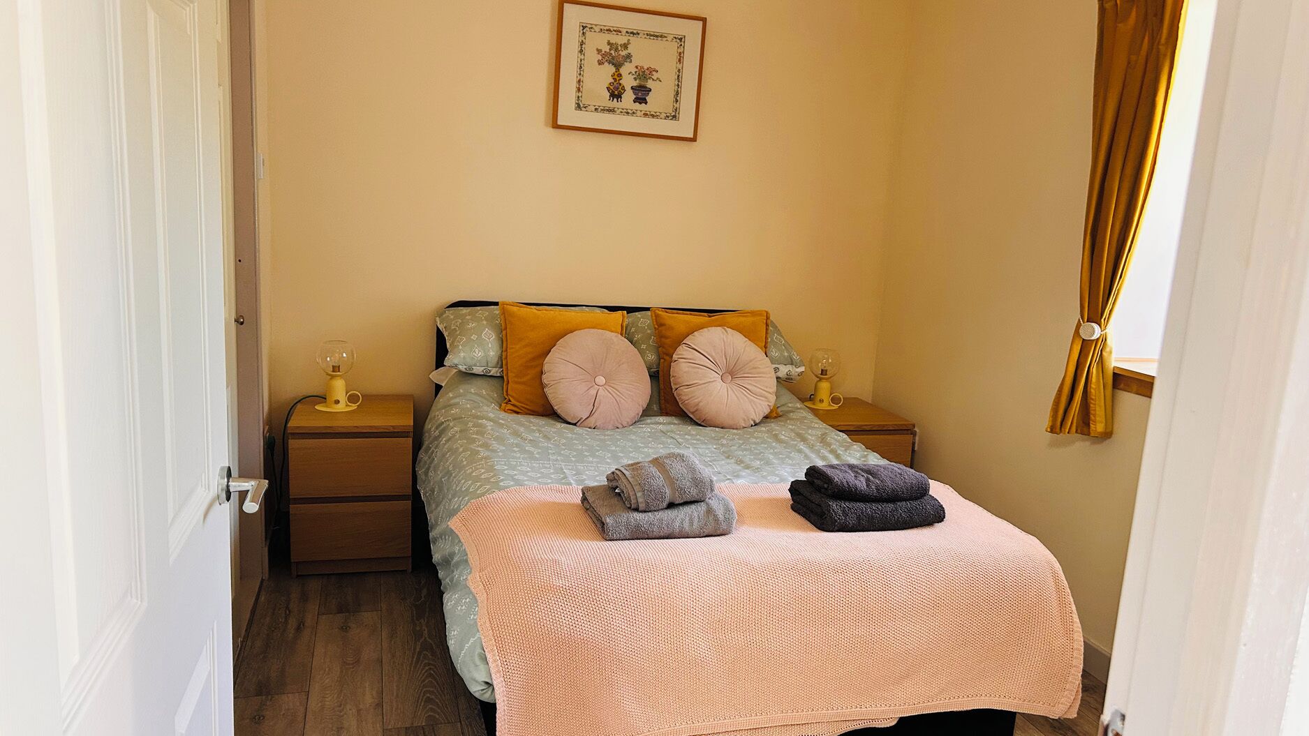 Un dormitorio ordenado con cama, dos mesitas de noche, un cuadro en la pared y una ventana con cortinas.