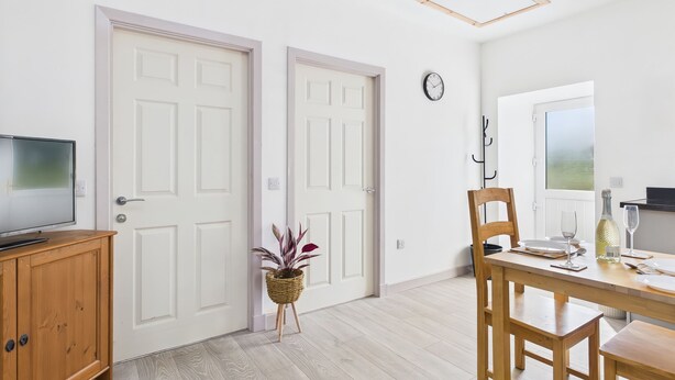 A modern room with two white doors, a wooden cabinet, a dining table with chairs, a potted plant, and a window with a view. - Bathach Beag (Oban)