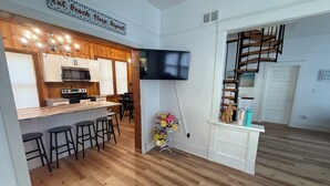 Una cocina moderna con detalles de madera, un comedor con taburetes de bar, un televisor en la pared y una escalera que sube a un nivel superior.