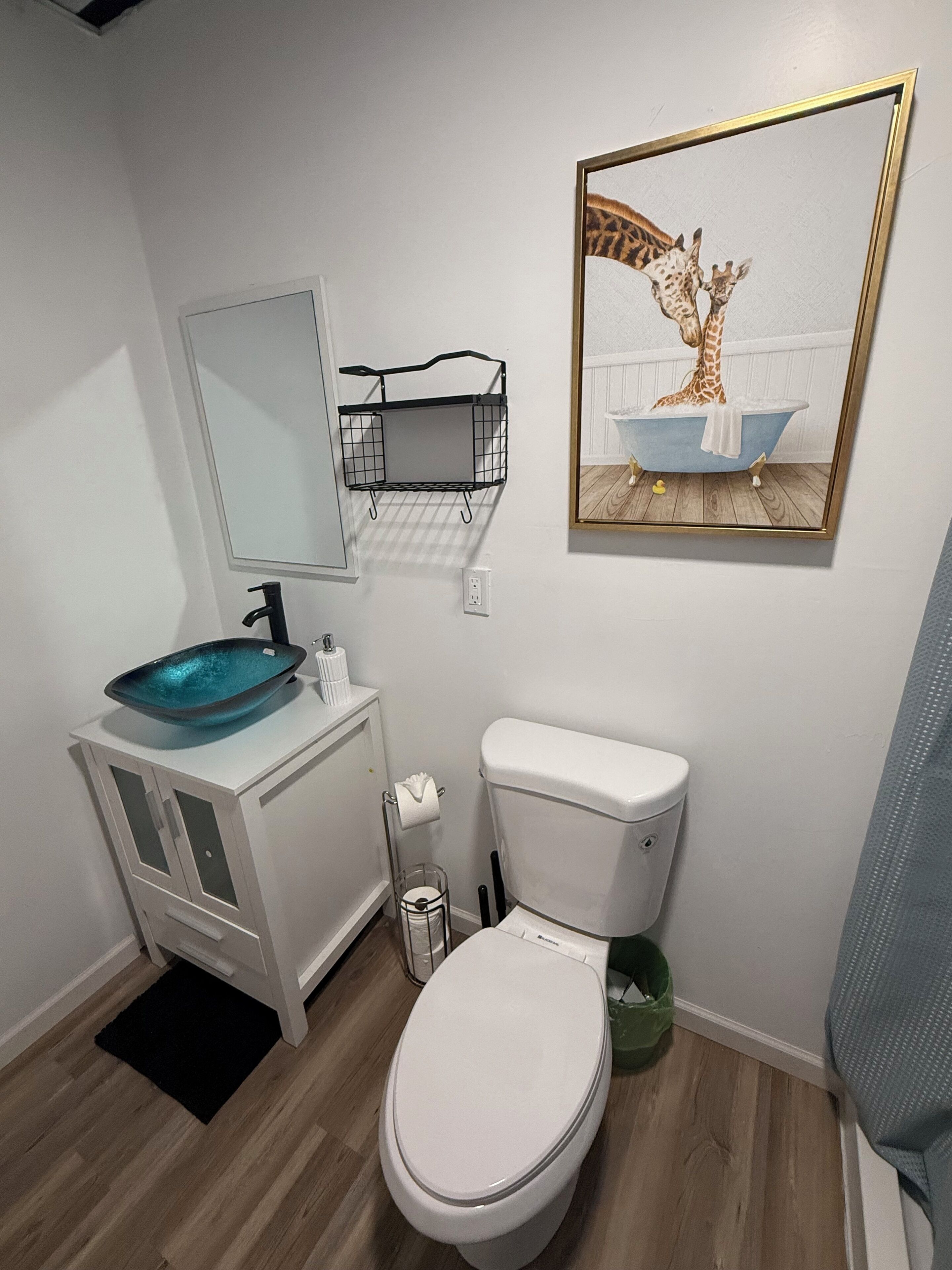 Un baño moderno con inodoro blanco, un lavabo tipo cuenco azul, un espejo y un cuadro con una jirafa en una bañera.