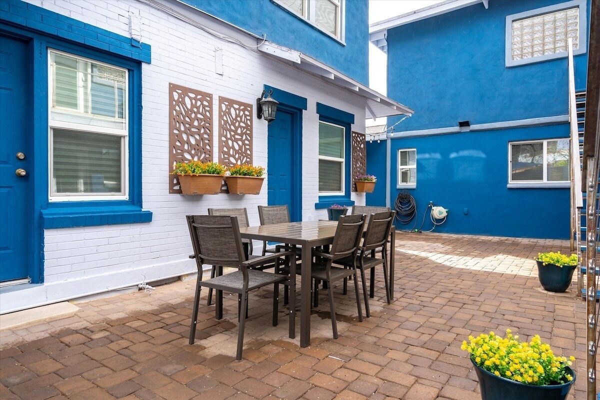 Une terrasse avec une table et des chaises, un bâtiment aux couleurs bleu et blanc, et des jardinières suspendues.