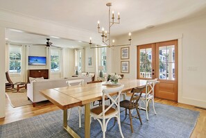 A dining area with a wooden table, white chairs, a chandelier, and a television in the background. - Old Village Luxury – Historic 5BR, Walk to Dining, Shops, & Sunset Views (Mount Pleasant)