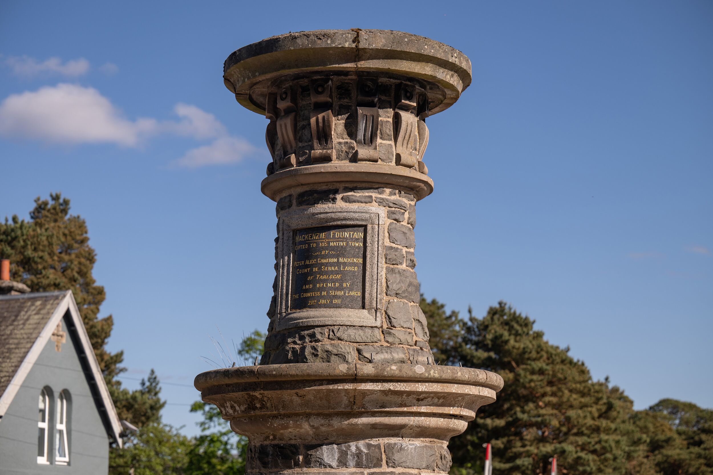 Ein Steindenkmal mit Inschriften, umgeben von Bäumen und einem Haus im Hintergrund.