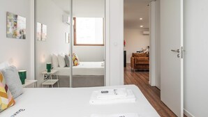 A modern hotel room with a bed, a small table with a lamp, a chair, and a sliding door leading to a dining area. - Liiiving in Matosinhos - Warm Vintage Apartment (Matosinhos)