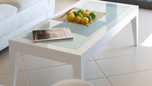 A modern living room with a white sofa, a glass coffee table with a magazine and fruit, and a view through a glass door to an outdoor area. - La Casa Rosa Seaside Getaway (Dorida)