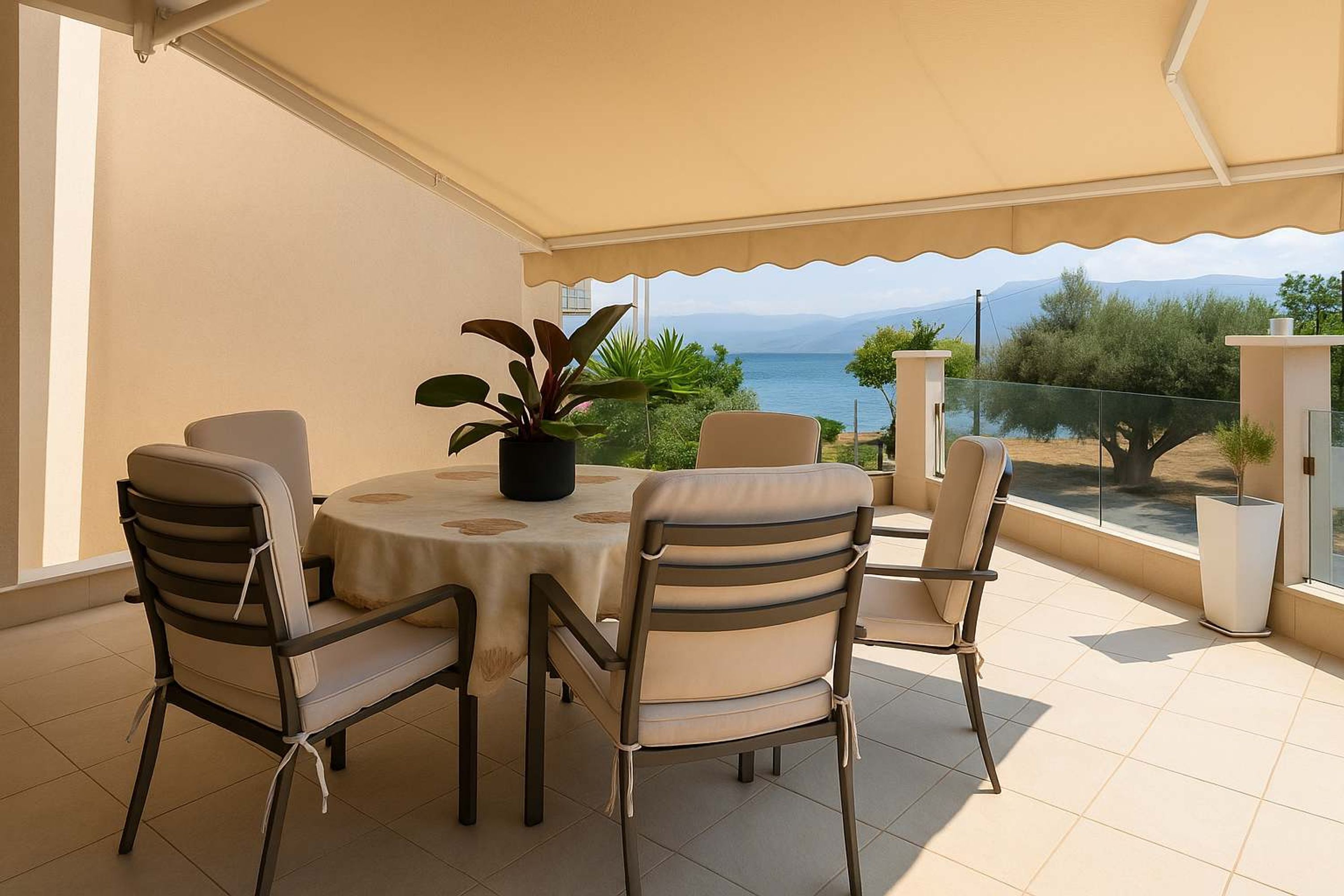 A covered outdoor seating area with a table and chairs, a potted plant, and a view of the sea and mountains.