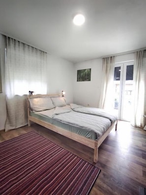A single-bed room with a wooden frame, a red striped rug, a painting on the wall, and a window with curtains. - Aria Plitvice (Plitvicka Jezera)