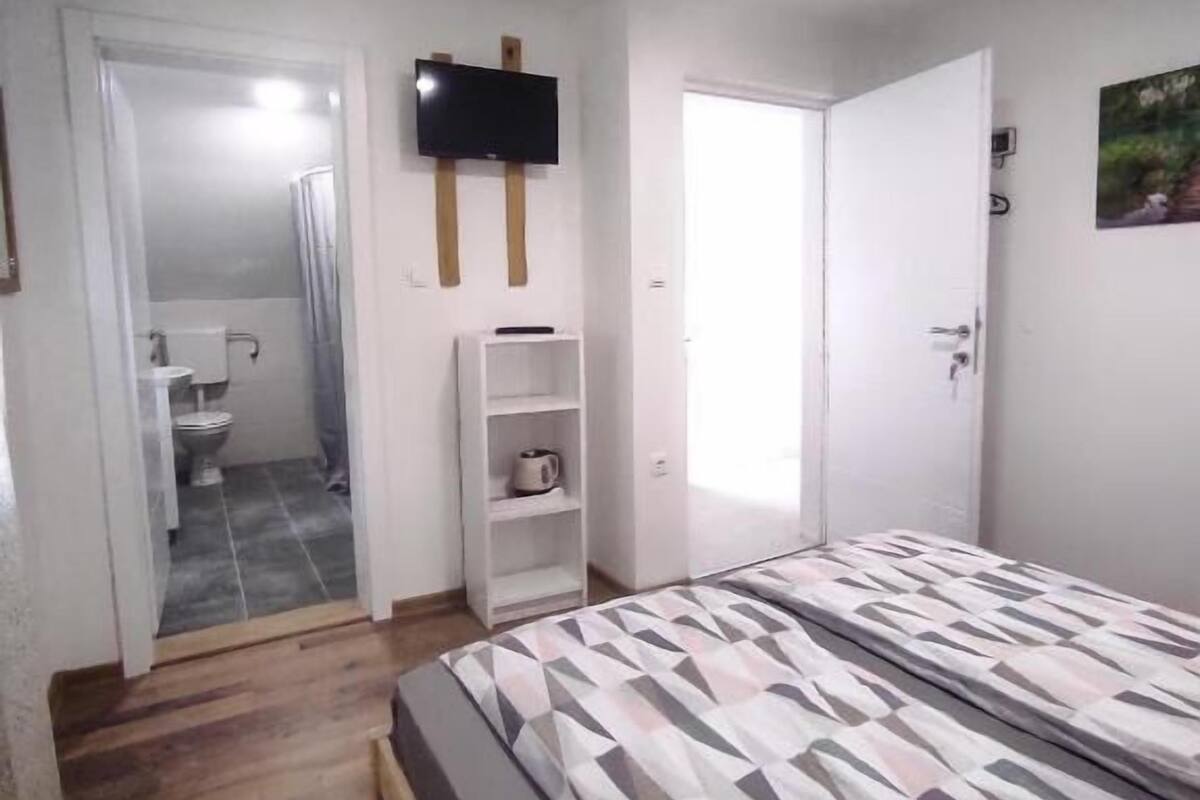 A bedroom with a bed, a TV mounted on the wall, a door leading to a bathroom, and a shelf with a cup.