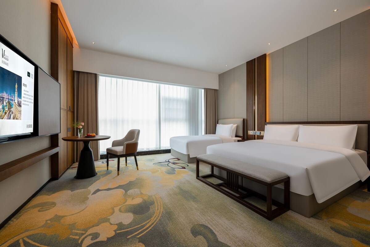 A hotel room with two beds, a TV, a small table with a vase, and a chair.