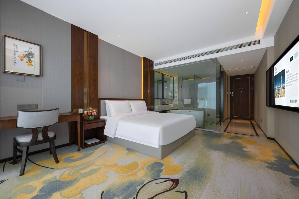 A modern hotel room with a bed, desk, chair, and a large screen.