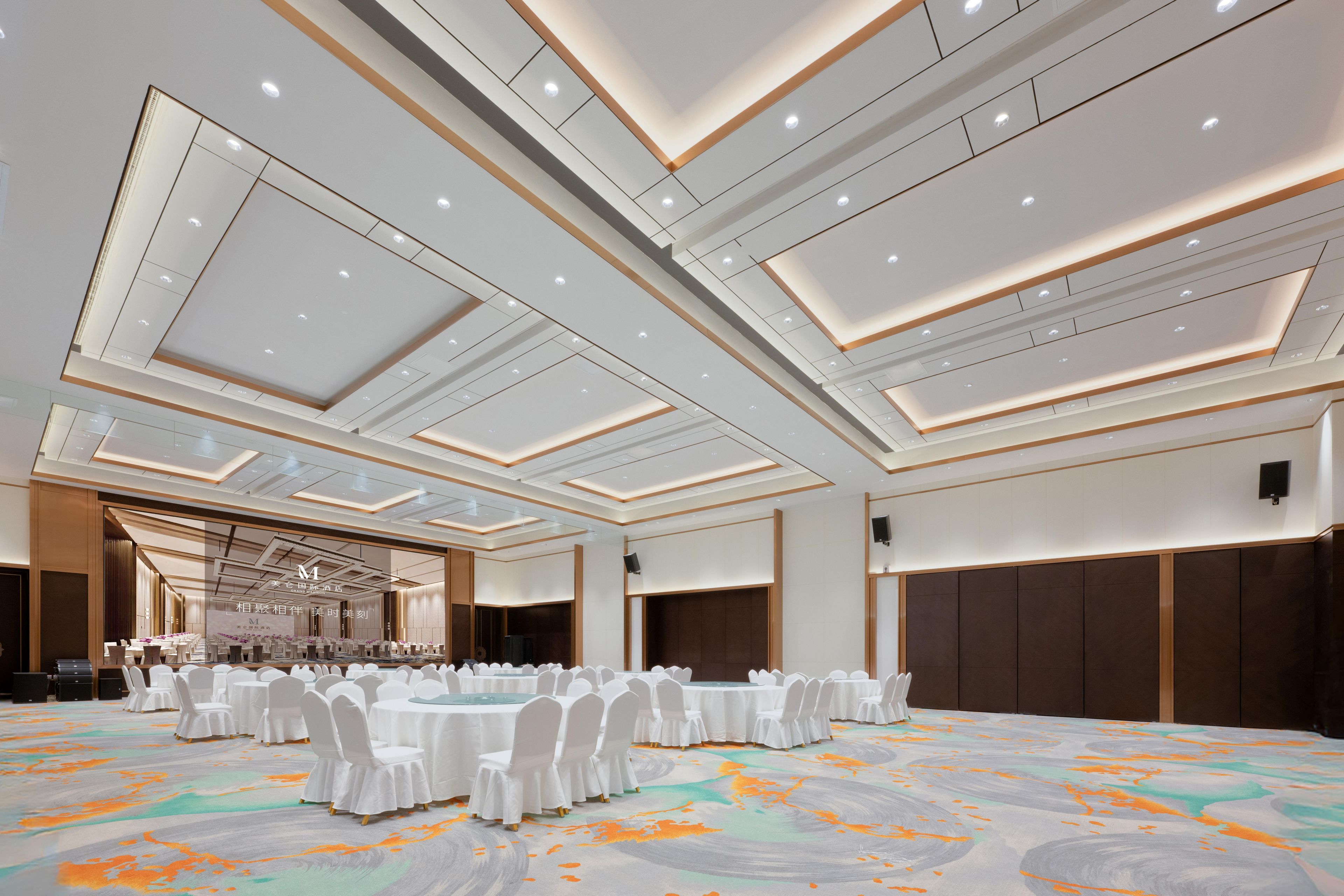 Un amplio salón de banquetes con mesas redondas, sillas blancas y una alfombra estampada.