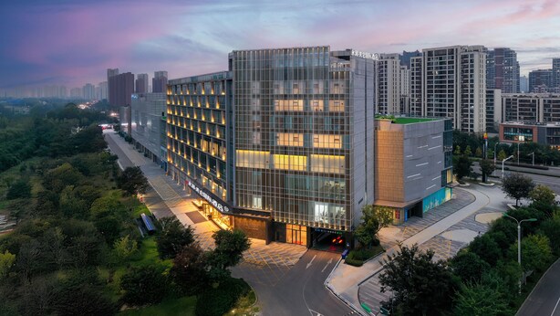 A modern hotel with a glass facade, surrounded by greenery and adjacent to a road.