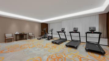Habitación de hotel con alfombra estampada, equipo de ejercicio, un escritorio con sillas y un ventanal con cortinas.
