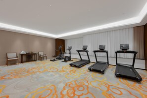 A hotel room with a patterned carpet, exercise equipment, a desk with chairs, and a large window with curtains.