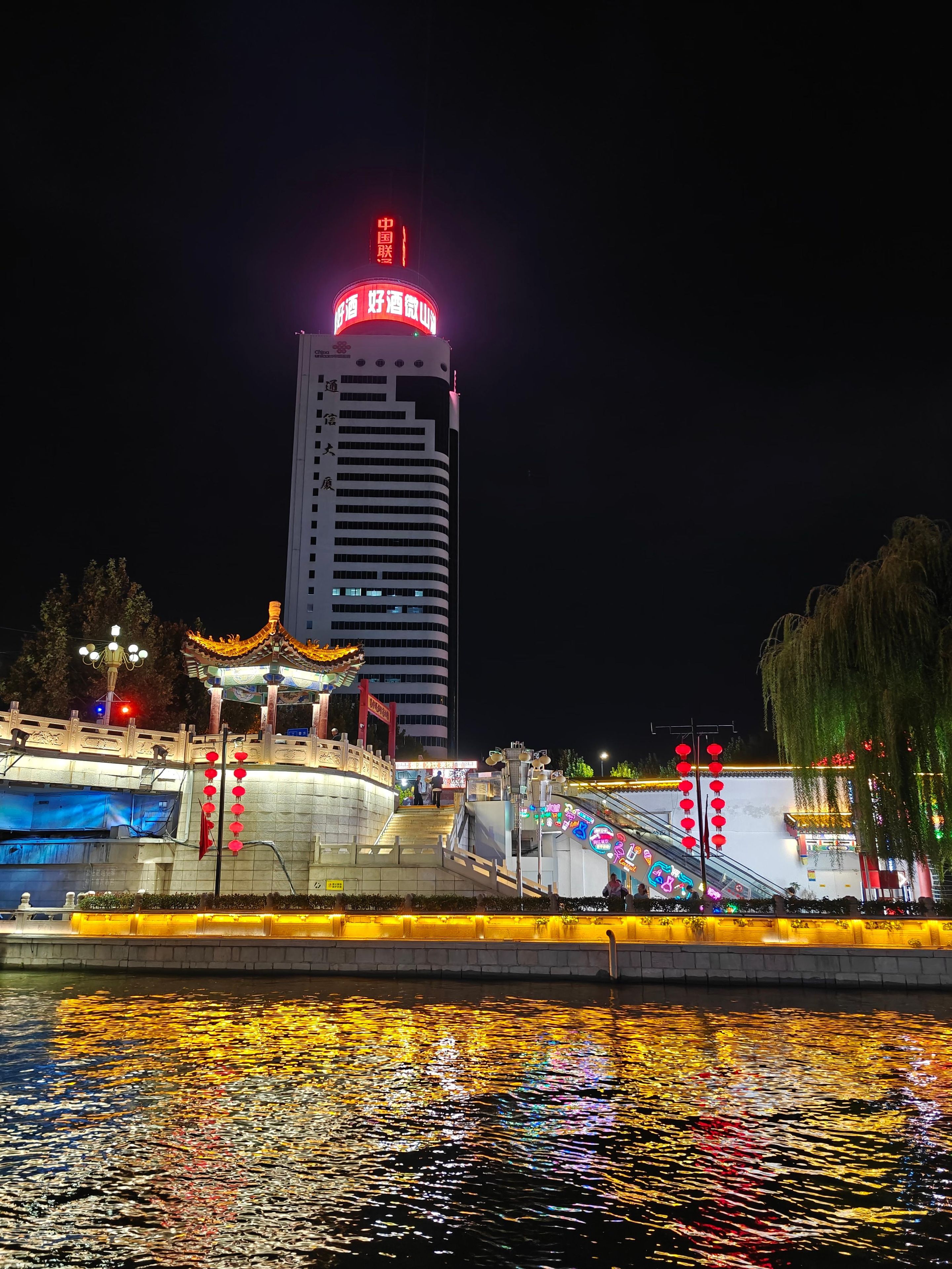 Vue nocturne d’un bâtiment moderne dont les enseignes sont illuminées, d’un pavillon traditionnel et d’une étendue d’eau qui reflète les lumières.