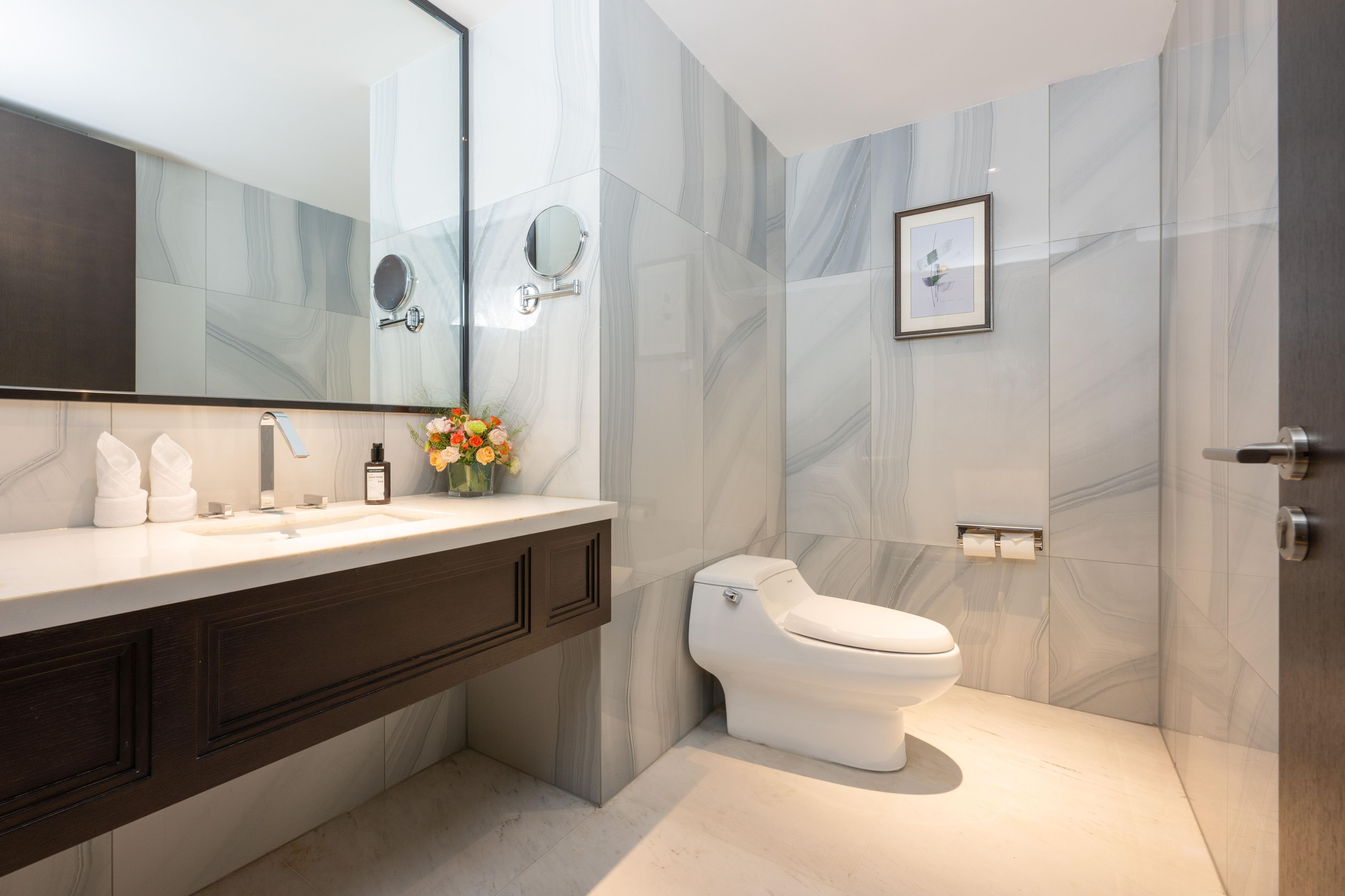 A modern bathroom with a marble wall, a white toilet, a large mirror, and a vanity with a sink and towel rack.