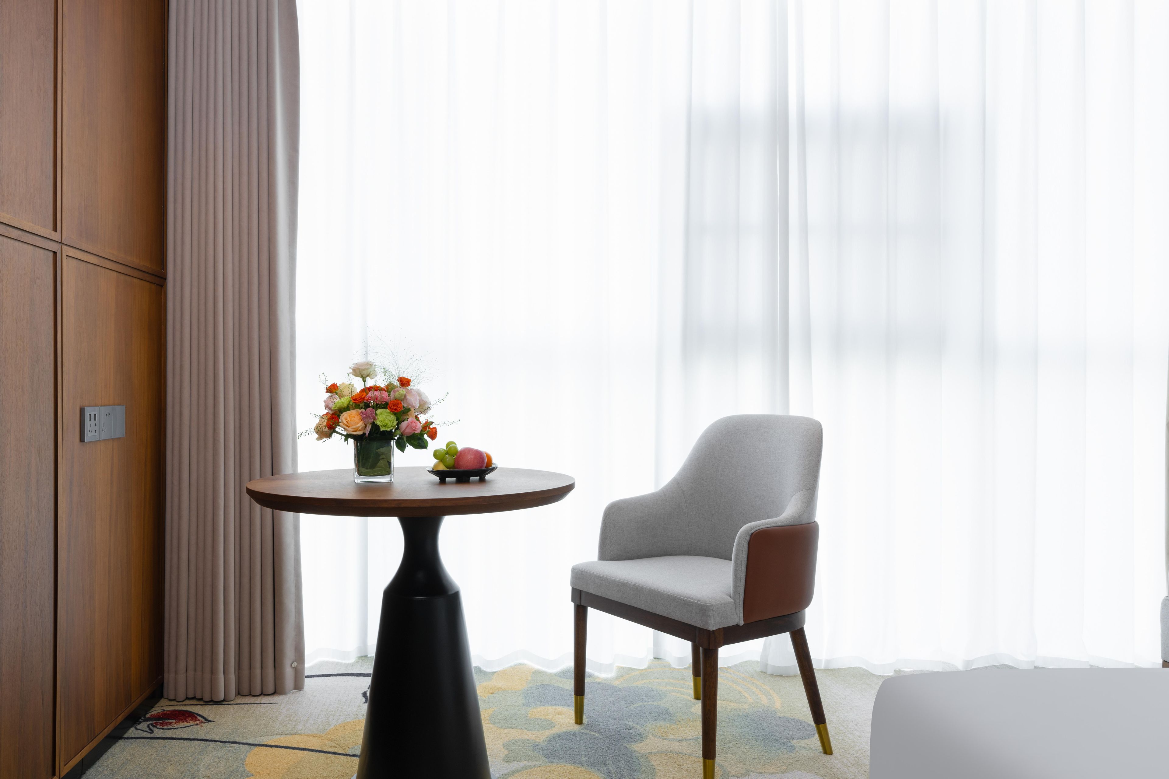 Habitación de hotel con pared de paneles de madera, una mesa redonda con un florero, un sillón gris y un ventanal con cortinas translúcidas.