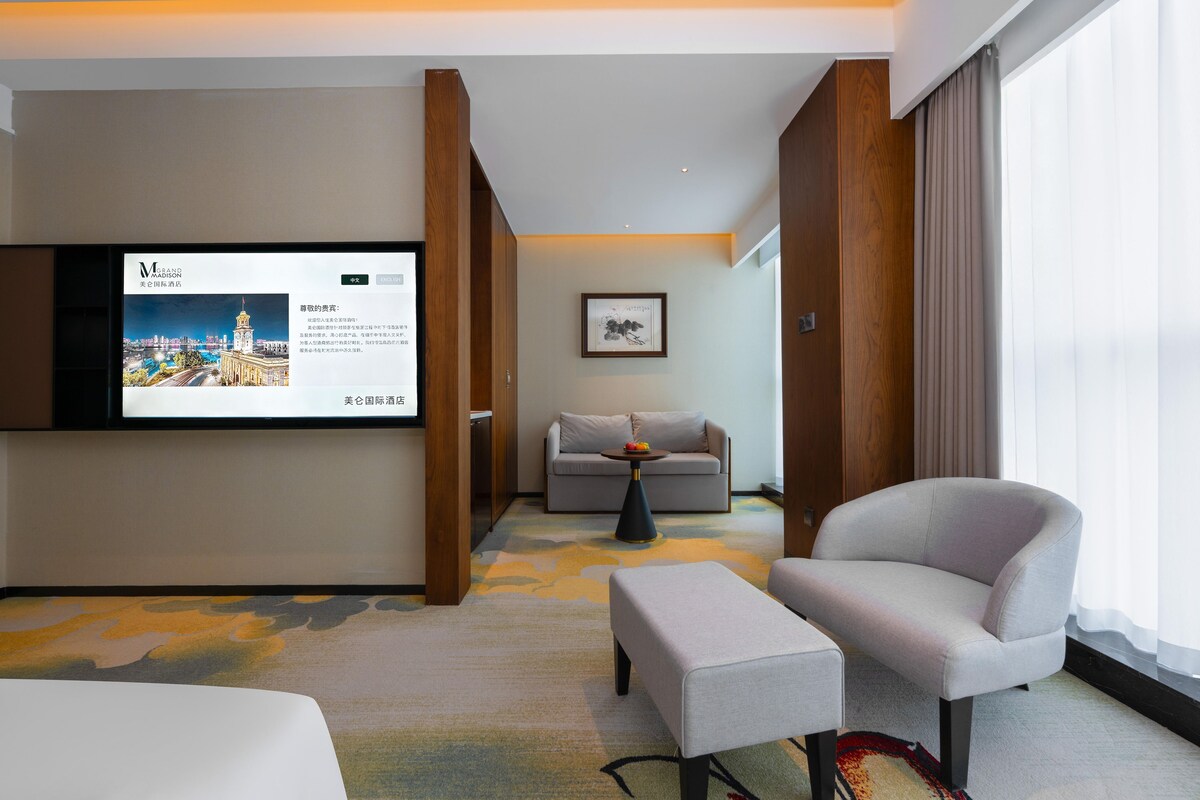 A hotel room with a bed, a sofa, a chair, a small table with fruit, a television displaying hotel information, and a framed artwork on the wall.