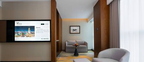 A hotel room with a bed, a sofa, a chair, a small table with fruit, a television displaying hotel information, and a framed artwork on the wall.