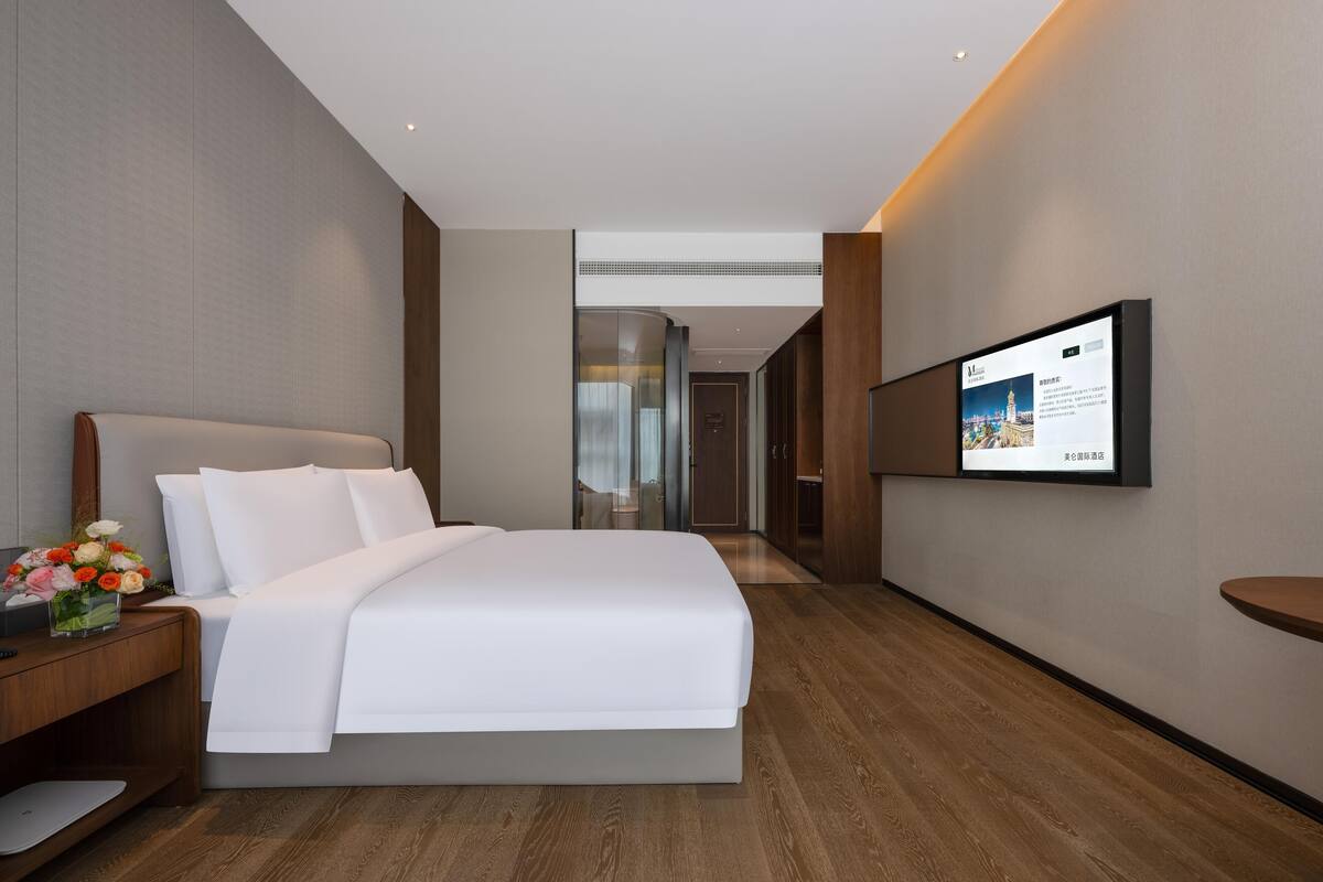 A modern hotel room with a large bed, a bedside table with a vase of flowers, a flat-screen TV mounted on the wall, and a wooden floor.