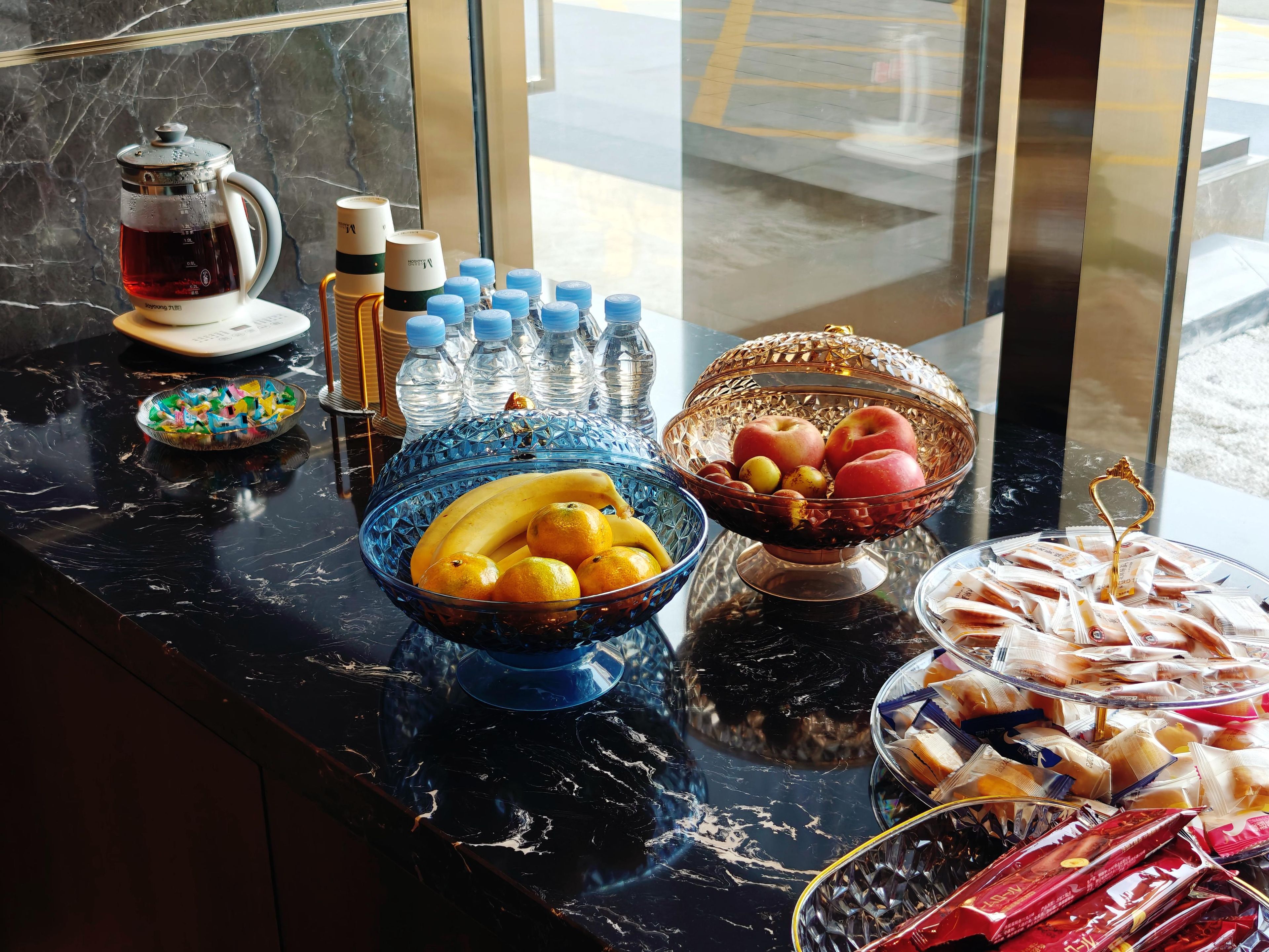 A buffet table with various food items and beverages, including a coffee pot, bottles, and bowls of fruits and pastries.
