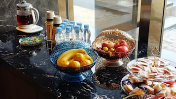 Una mesa de buffet con diversos alimentos y bebidas, incluyendo una cafetera, botellas y cuencos con frutas y pasteles.
