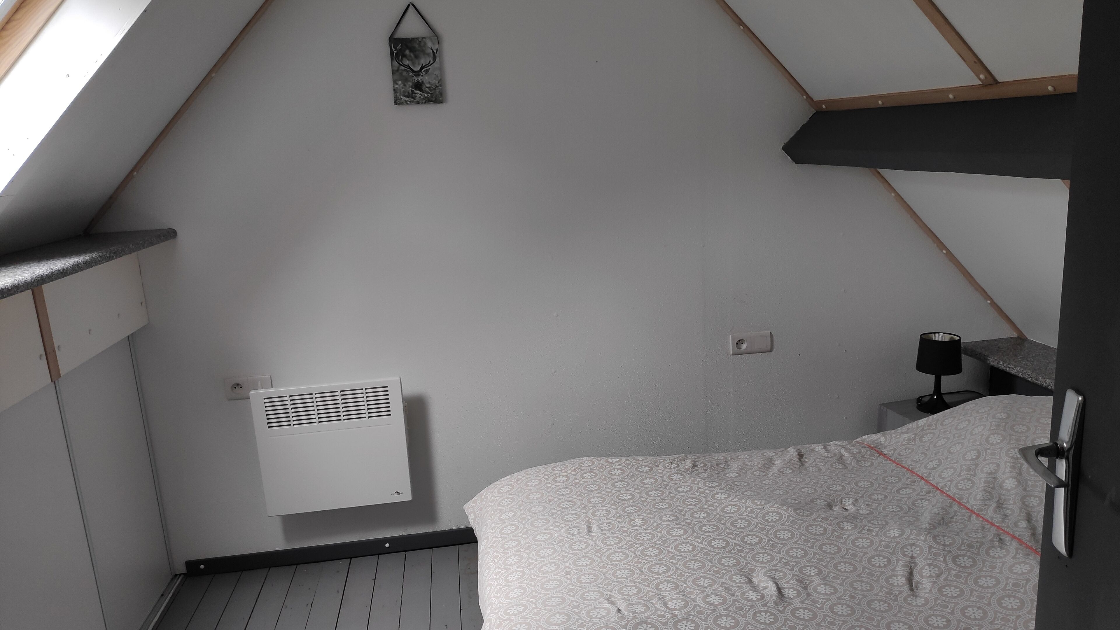 A bedroom with a sloped ceiling, a bed with a patterned bedspread, a bedside table with a lamp, and a wall-mounted heater.