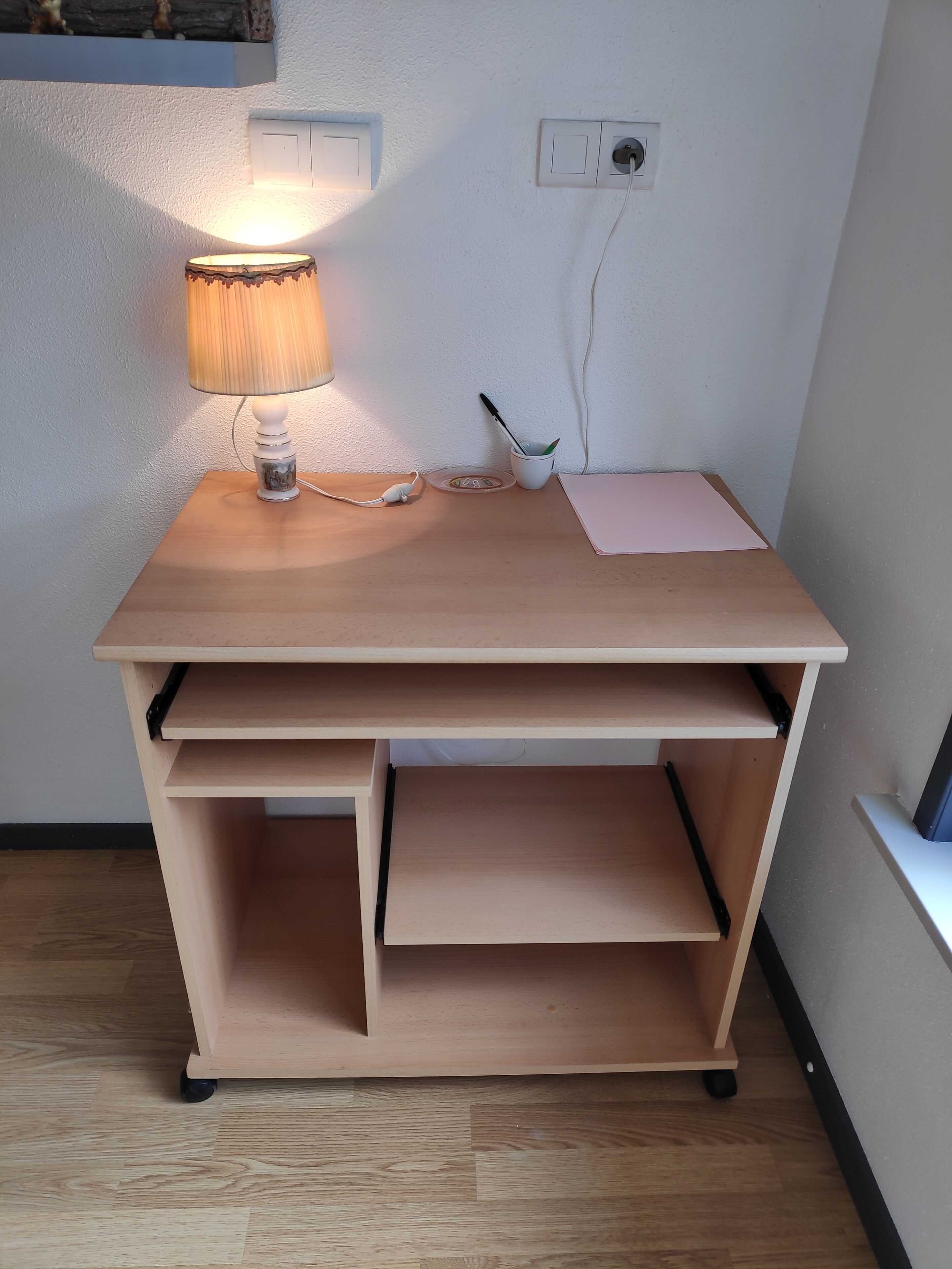 A wooden desk with a lamp, a power outlet, and a cup with a spoon.