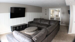 A living room with a large sectional sofa, a flat-screen TV mounted on the wall, and a door leading to another room. - Spacious 5-bedroom house in charming Stansbury Park with WiFi, AC (Stansbury Park)