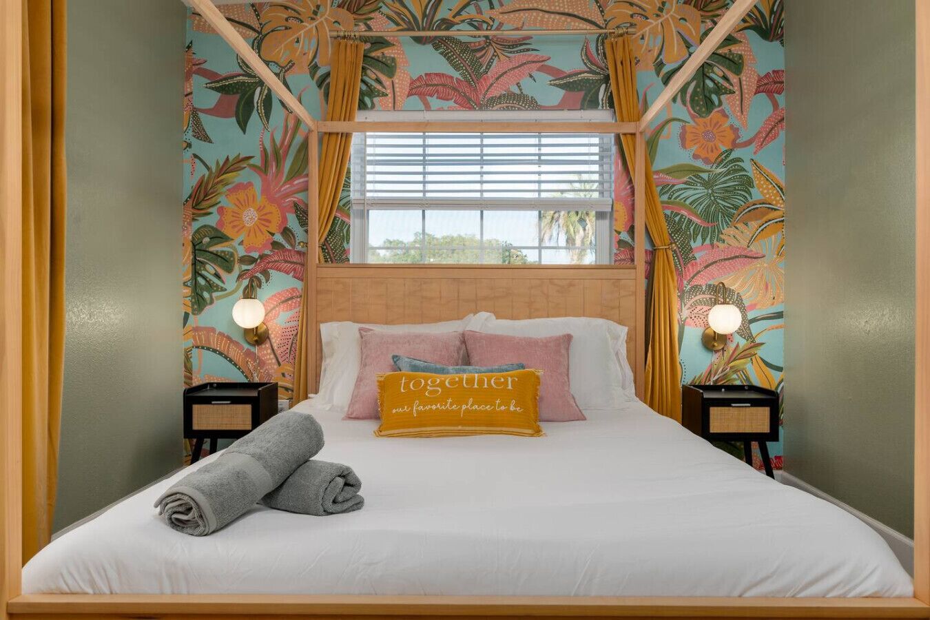Une chambre à coucher avec un lit, des oreillers, une serviette enroulée, une fenêtre avec des stores et un mur orné d’un motif floral.