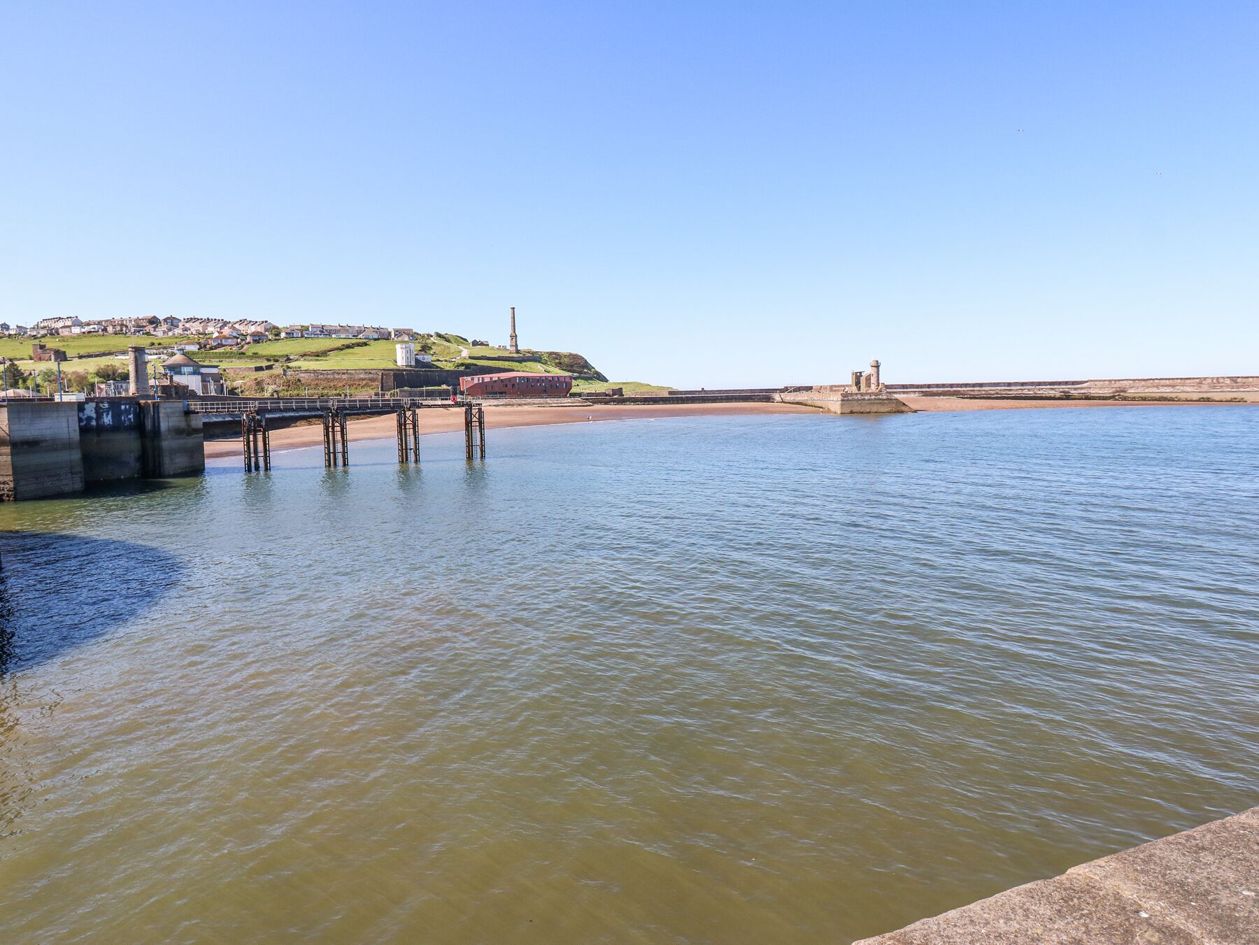Eine Küstenlandschaft mit einem Pier, der sich in ruhiges Wasser erstreckt, einem Leuchtturm auf einem Hügel und einer in der Ferne liegenden Stadt.