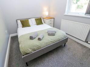 Un lit bien fait, recouvert d’une couverture verte, avec deux serviettes enroulées et des oreillers, dans une pièce dotée d’une fenêtre et d’un radiateur.